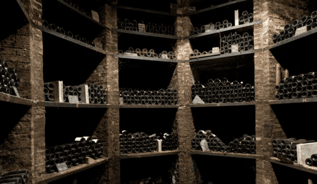 A dimly lit, historic wine cellar at Domaine des Comtes Lafon, showcasing rows of aging wine bottles stacked on rustic stone shelves, preserving decades of Burgundy winemaking tradition.