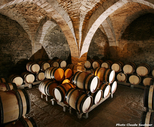 A photo of Domaine Dugat-Py wine cellar, taken by Claude Saulnier. The picture shoes many barrels of Burgundy Wine, kept in a classic stoned cellar where the temperature is cool, perfect for wine.